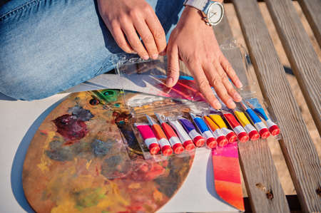 Close-up of female artist hands on a painting palette and tubes with paints on a wooden surfaceの写真素材