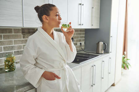 A beautiful woman in a white robe, leading a healthy lifestyle, drinking lemon water leaning against...