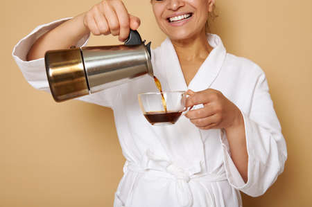 Smiling mature woman with toothy smile in white bathrobe pouring coffee drink from a geyser coffee...