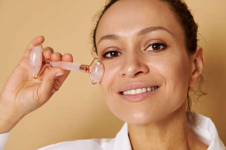 Head shot of woman with beautiful toothy smile massaging face with jade roller massager for face...