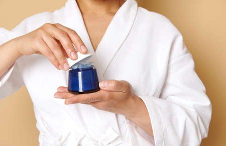 Close-up of a blue bottle with facial moisturizer in the hands of a woman in white waffle bathrobe. Isolated over beige background with copy spaceの写真素材