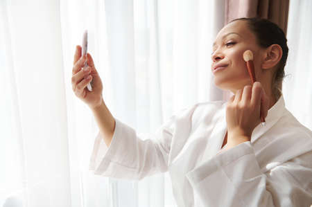 Beauty portrait of an attractive mixed race woman standing in front of sunlight by the window and looking at her reflection in the mirror while applying blush or highlighter to her faceの写真素材