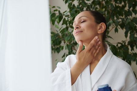 Hydrating and moisturizing. Young African American woman in white bathrobe caring of her skin, applying face moisturizer , tenderly massaging her neckの写真素材