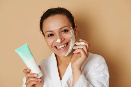 Close-up beauty portrait of a young woman with beautiful toothy smile applying a facial cream or...