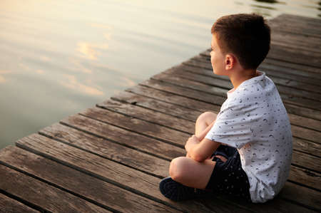 Serene schoolboy sitting on a pier and enjoying the tranquility of nature scene while looking at the...