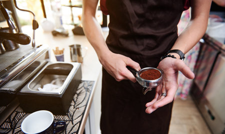 Partial view of unrecognizable barista in apron holding coffee press and portafilter with ground coffeeの写真素材