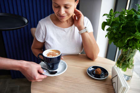 Young African American woman in a cafe looking at a cup of cappuccino in the hands of a waitress serving herの写真素材