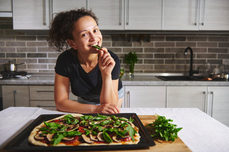 Smiling African American woman leaning on kitchen table with hot pizza and tasting basil, smiling,...