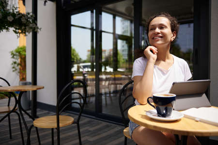 Coffee break in open air. Smiling brunette in white t-shirt and jeans shorts looking away, relaxing sitting at a table with coffee cup in summer terrace of cafeteria.の写真素材