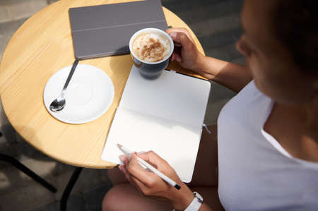 High angle view of a woman writing on a notebook sitting at a table with a coffee in her hand. Freelance, business and outdoor work conceptsの写真素材