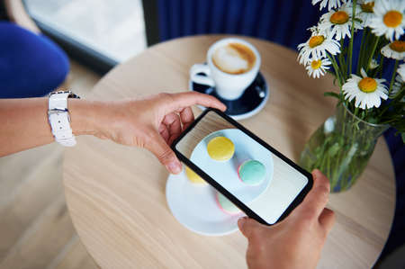 Close-up of woman hands taking a photo of plate with macarons on a wooden table in cafeteria. Coffee break time. Mobile phone in live view modeの写真素材