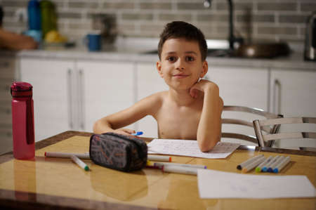 Adorable boy looking at the camera while sitting at the table and drawing at home