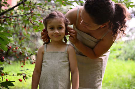 Enjoying time together. Happy family. Positive human emotions. Mom and daughter having a lovely time together during the cherry harvest. Mom puts cherry berry earrings on her daughter's ears.の写真素材