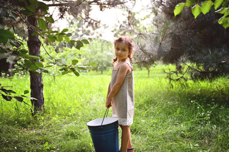 Beautiful little girl in a linen dress stands in the garden with a large blue metal bucket in her hands at sunsetの写真素材