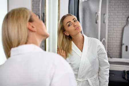A gorgeous woman in a white robe touches her blonde hair, admiring her reflection in the mirror. Beauty portrait of attractive woman with perfect smooth facial featuresの写真素材