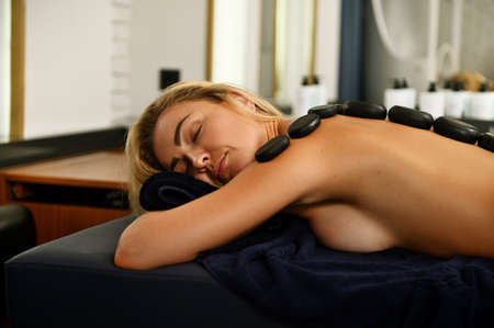 Calm pretty European woman relaxing with closed eyes on massage table during hot stone massage at...