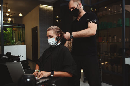 Handsome professional hairdresser in black outfit combing hair of business woman, client visiting beauty salon and working remotely on laptopの写真素材