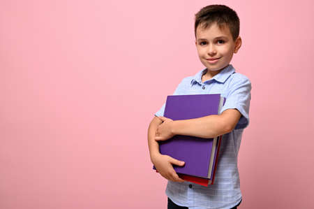 An adorable schoolboy holds books in his hands a cute smiles posing on camera, isolated over pink...