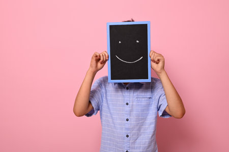 Unrecognizable school boy covers his face with a chalkboard with drawn smiling emoticons, expressing happiness. Isolated over pink background with copy spaceの写真素材