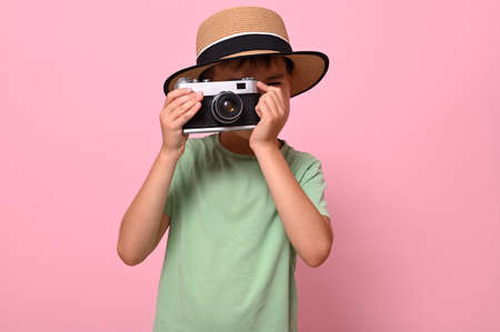 Cute schoolboy in green t-shirt and summer hat taking photo using an old vintage retro camera, isolated over pink background with copy spaceの写真素材