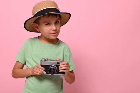 Handsome boy in summer hat holding a vintage retro camera, looks at the camera posing against pink background with space for textの写真素材