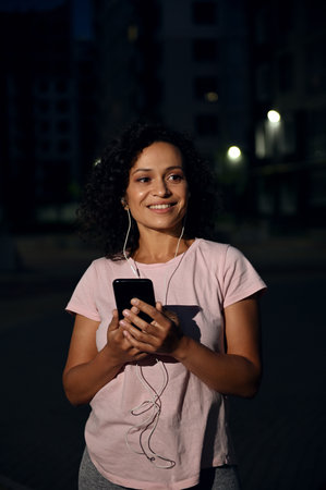 Portrait of African American woman with earphones and smartphone, smiling with toothy smile, standing against illuminated buildings backgroundの写真素材