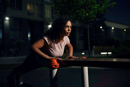 Young mixed race woman athlete performing push-ups from a wooden bench on the sports ground and pumping chest and shoulder muscles during evening workoutの写真素材