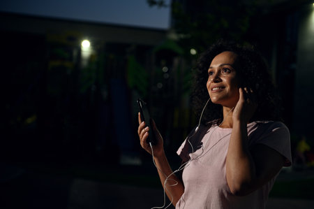 Side portrait of a Hispanic sportswoman with earphones, holding a mobile phone and enjoying...