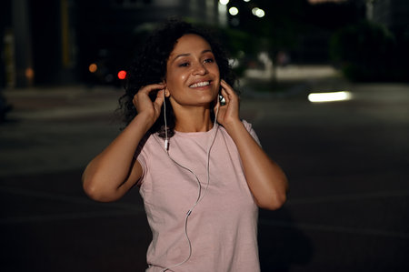 Portrait of cute young Latin American woman wearing headphones, smiling looking to the side standing against evening lights backgroundの写真素材