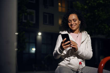Smiling mixed race woman in sportswear sitting on the wooden bench wearing earphones and using...