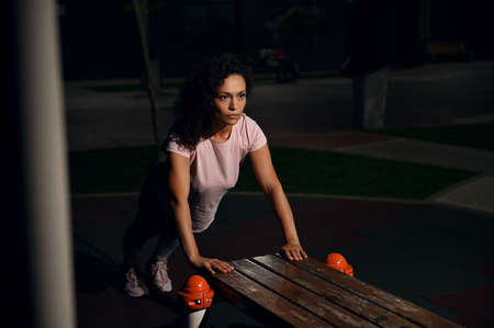 Young female athlete of mixed nationality doing push-ups from a wooden bench on the sports ground...