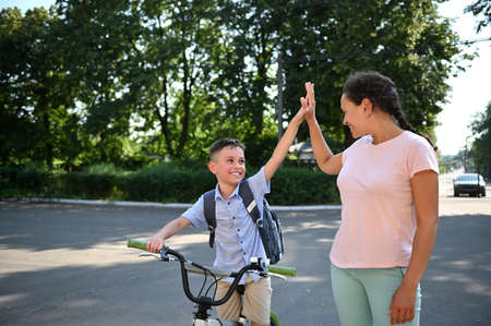 Smiling school boy on bicycle giving high five to his African American mother. Child coming back to school. Kids ridingの写真素材