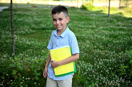 Happy elementary student coming back to school. Adorable schoolboy holding a workbooks and smiling to camera standing on green grass background.の写真素材