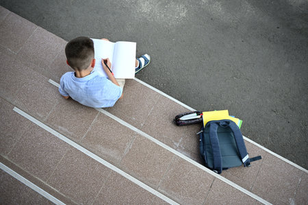 A schoolboy concentrated on doing homework, sitting on the stairs of school building next to his...