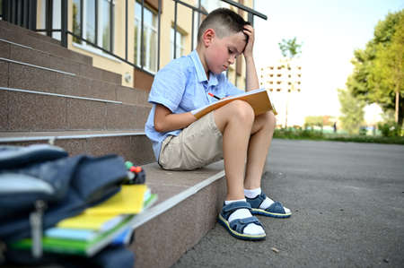 Schoolboy concentrated on doing homework, sitting on the stairs of school building, blurred backpack with workbooks on the foreground. Back to school concept.の写真素材
