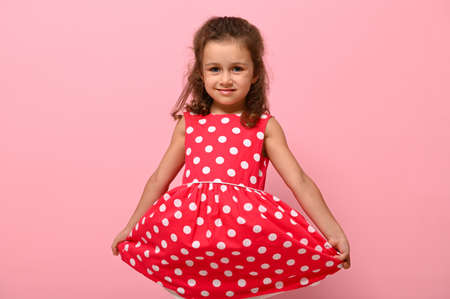 Portrait of cute charming girl in a pink dress with white polka dots posing onpink background cute smiling and holding the bottom of the dress with her hands. Beautiful child portrait for advertisingの写真素材