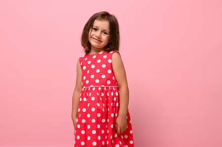 Gorgeous European baby girl, adorable child dressed in pink polka-dot dress, cute smiles posing over pink background with space for text.の写真素材