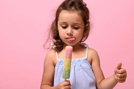 Headshot, close-up portrait of a 4 years cute baby girl with healthy vegan ice cream in hand and showing thumb up. Pink background, copy space . Summer dessert, cheerful summer mood conceptの写真素材