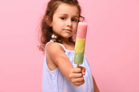 Focus on multi colored colorful frozen juice, ice cream on stick in hands of adorable girl, out of focus, isolated on pink background with copy spaceの写真素材