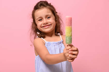 Funny cheerful little girl smiles showing to the camera a delicious sweet frozen juice, ice cream in her hands. Summer concept for advertisement , pink background, copy space for promotionの写真素材