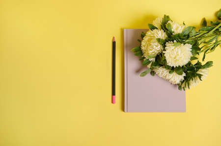 Flat lay of beautiful delicate bouquet of Autumn flowers, white asters lying down on the corner of a notepad organizer, next to a wooden color pencil, isolated over yellow background with copy spaceの写真素材
