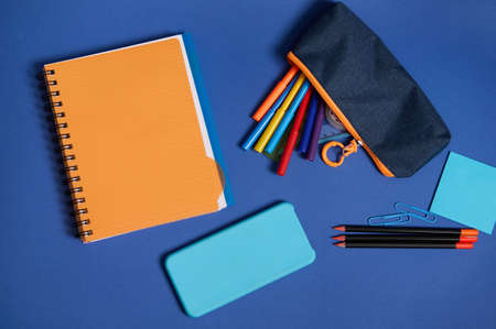 Top view of school office supplies and a smartphone lying screen down on a blue background . Flat lay composition of stationery in two contrasting colors, blue and orange with copy spaceの写真素材