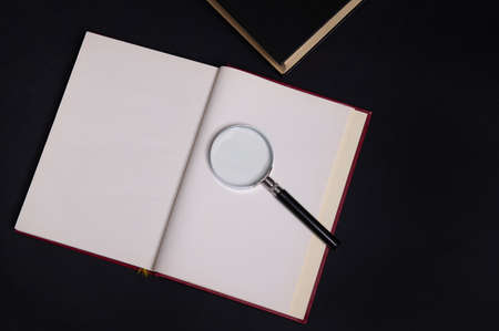 Flat lay of a magnifying glass, loupe on an opened book in hard red cover, isolated over black...