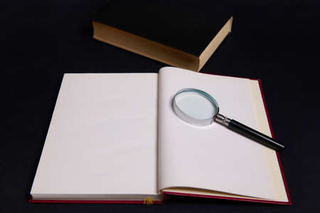 A magnifying glass, loupe, magnifier on an opened book in hard red cover, isolated over black...