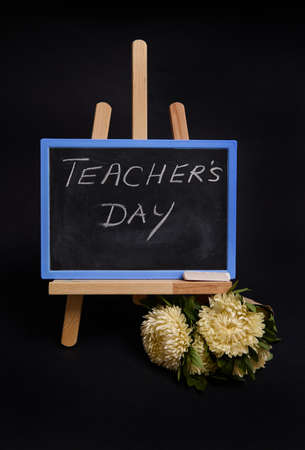 Close-up of a bouquet of asters flowers next to a chalk chalkboard with lettering Teachers Day, standing on a wooden table easel, isolated on black background with copy space.の写真素材