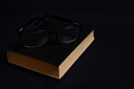 Monochrome composition of eyeglasses on a book in hard black cover, isolated over black background with space for text. Teacher's Day concept, Knowledge, literature ,reading, eruditionの写真素材