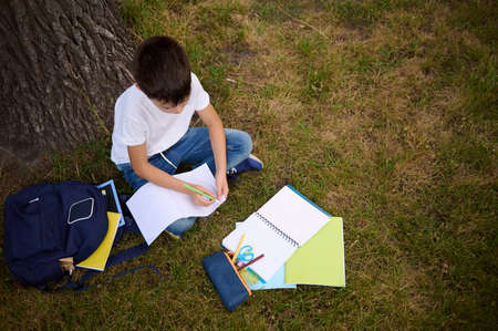 Top view of school boy focused on writing on empty blank sheets of a workbook, doing school tasks sitting on green grass at the park, after school . School accessories and backpack lying on grassの写真素材