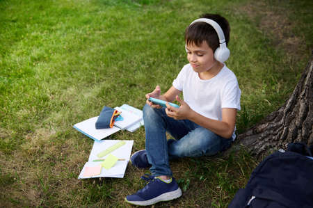 Top view of an adorable handsome elementary aged schoolboy wearing wireless headphones, focused on playing with smartphone, resting on the green grass of urban park, school supplies lying down next to himの写真素材