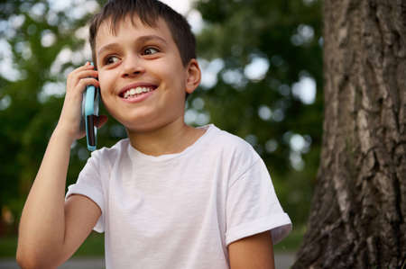 Cheerful handsome happy school child boy talking on mobile phone, cute smiling looking away, resting on the public park after first day at school on beautiful summer day. Concepts of Back to schoolの写真素材
