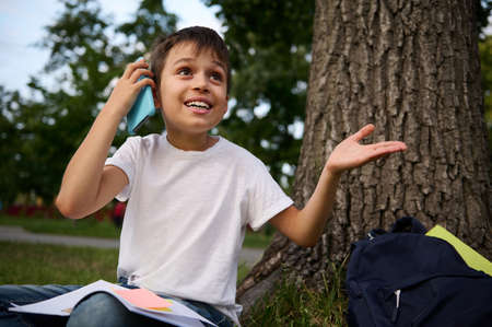 Handsome cheerful adorable school child boy talking on mobile phone, cute smiling looking away, gesturing with hands, resting on the public park after first day at school on beautiful summer day.の写真素材